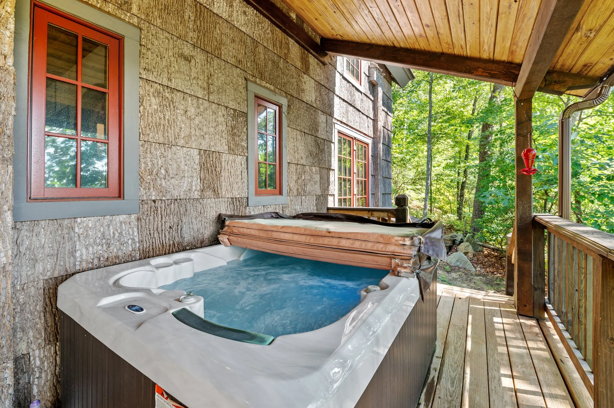 Outdoor hot tub on wooden deck surrounded by forest canopy with red window frames