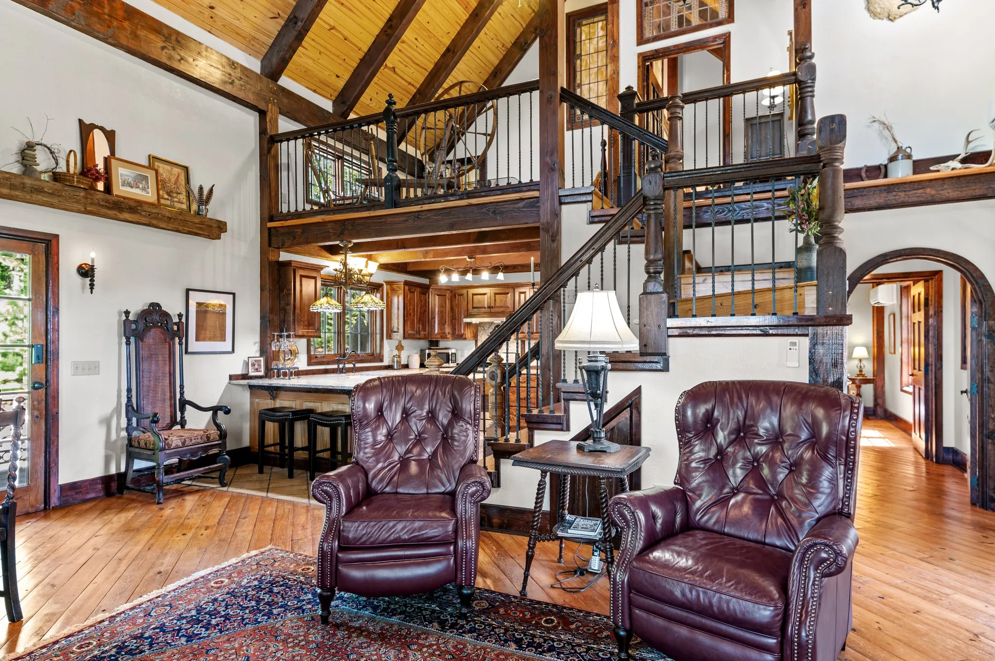 Open-concept living area with loft view, leather furniture, wooden beams, and iron railing
