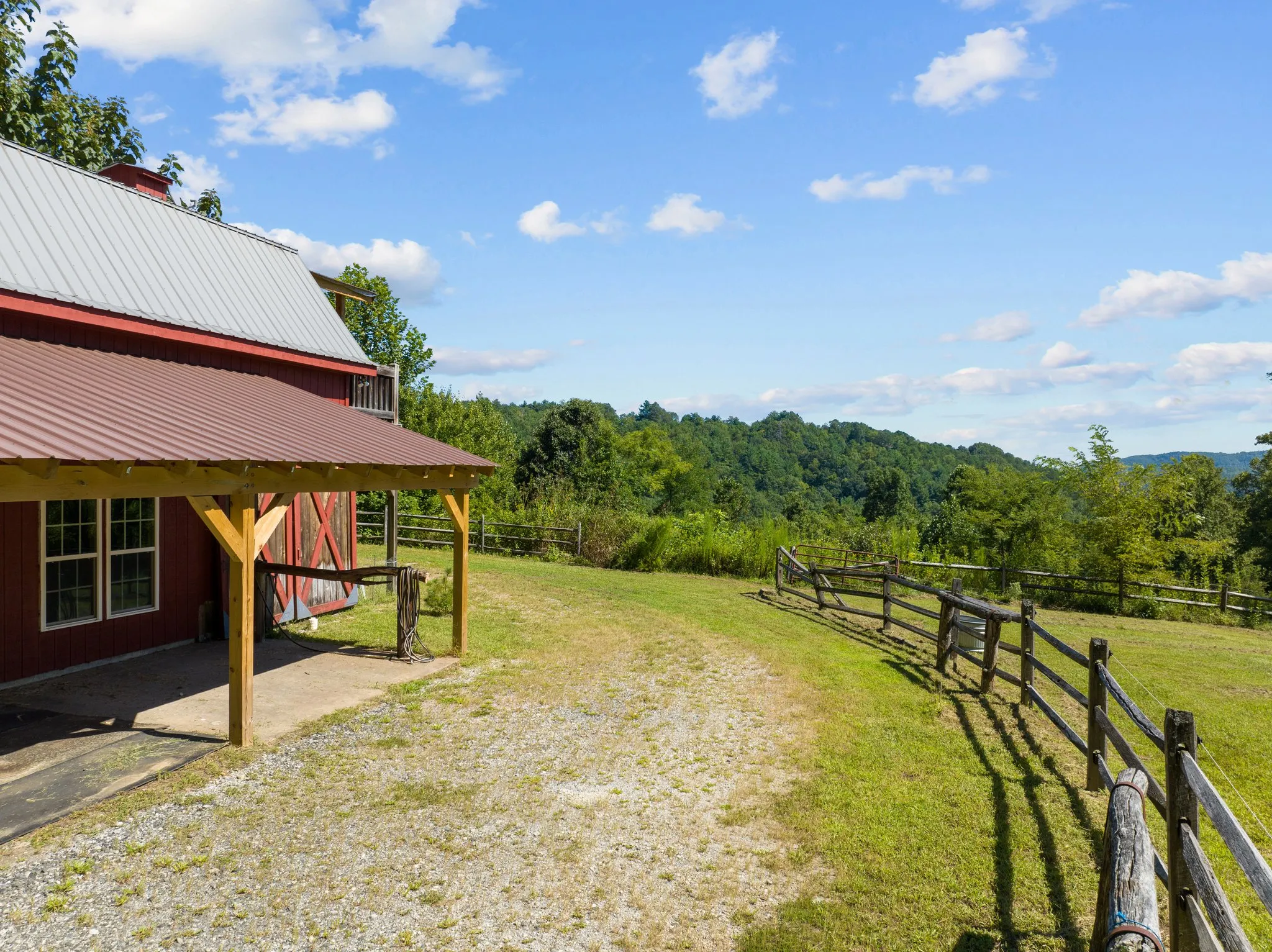 Red barn with covered porch and split rail fence overlooking green pasture and mountain forest