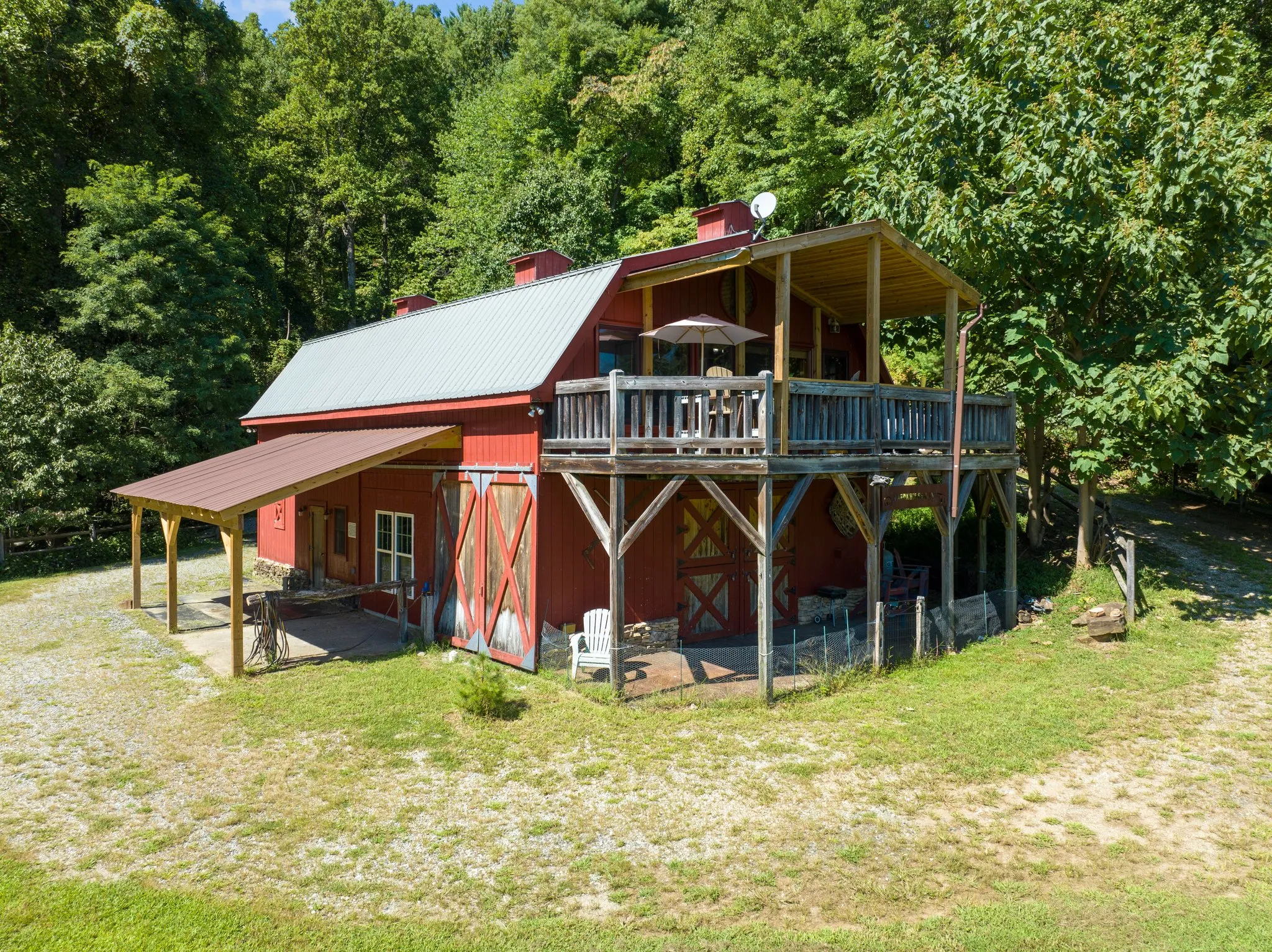 Two-story red gambrel barndominium with upper deck, covered porch, and surrounding forest clearing